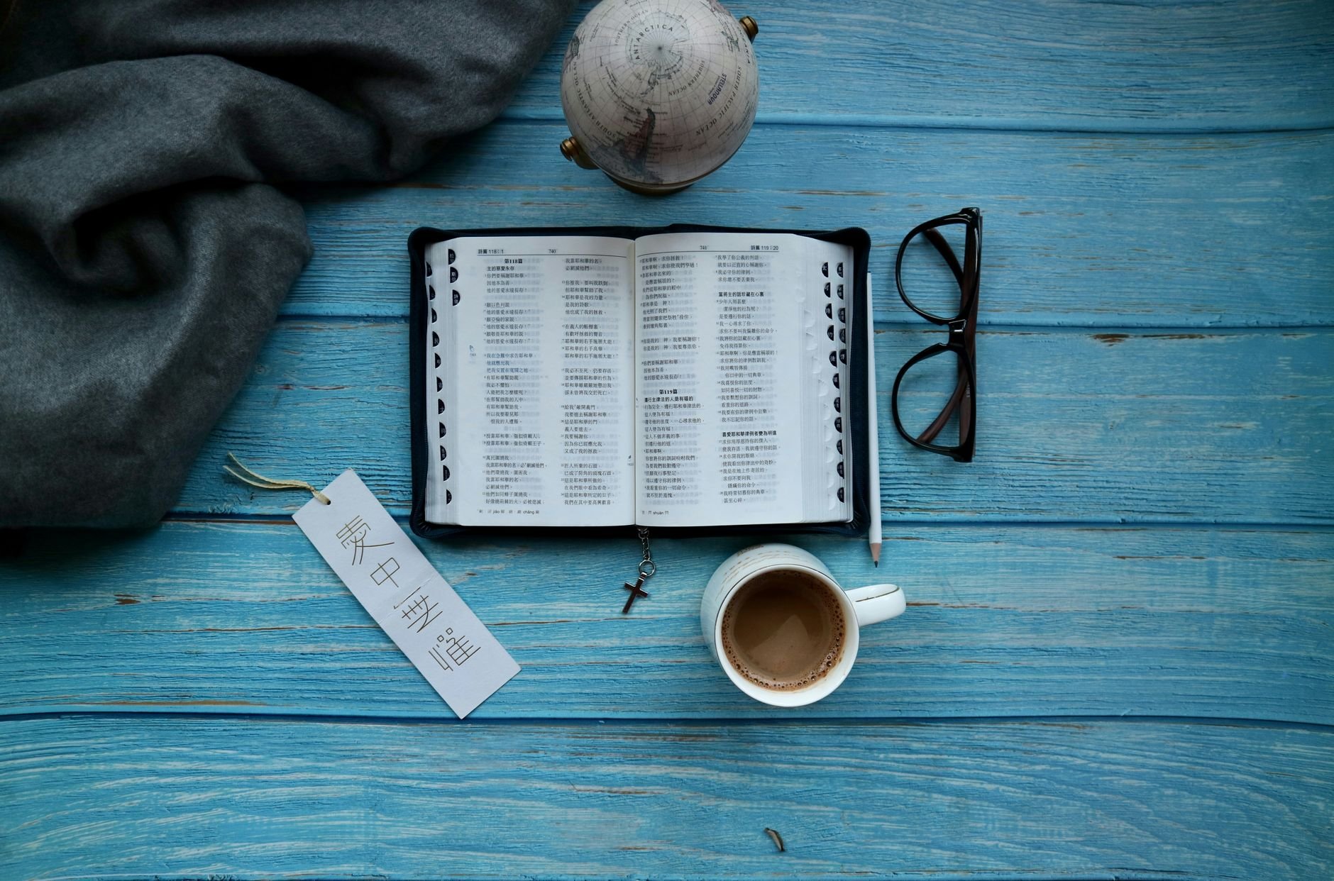 An open Bible with a bookmark, a coffee cup, glasses, and a globe on a table.